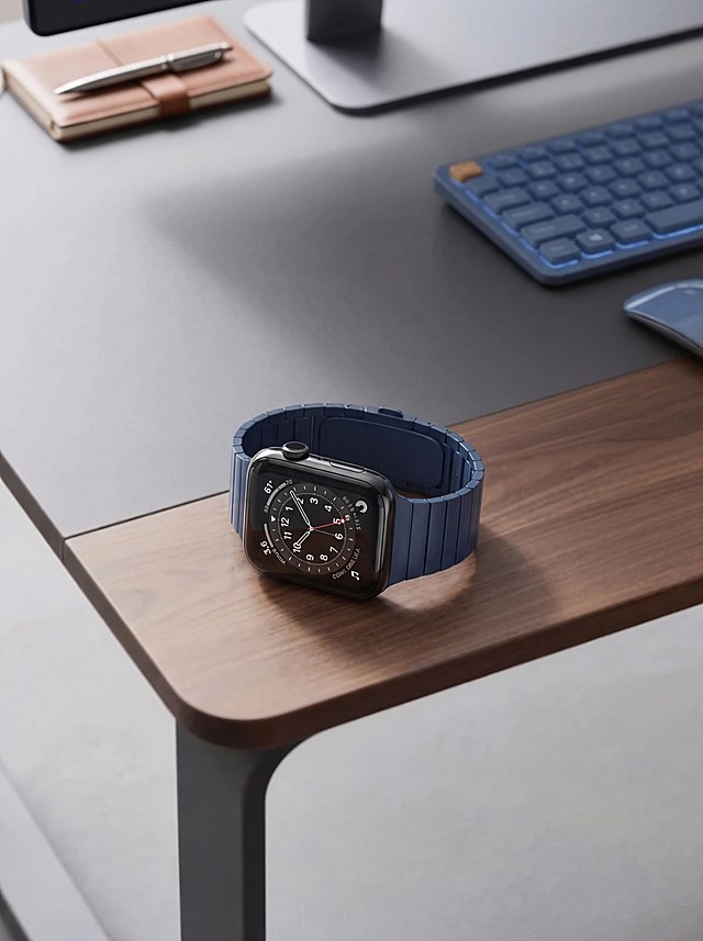将图二的智能手表放到图一的木质桌面上，保证手表物理细节和桌子背景图内容绝对不变。手表是黑色金属表壳搭配深蓝色表带，表盘清晰，整体设计简约精致。精准匹配原场景透视角度，实现空间尺度物理融合，确保手表自然放置在桌面。重置手表光影，增强光照一致性，赋予自然光影及真实接触阴影。手表色温亮度与环境完全协调，金属和玻璃材质增加环境映射。消除视觉接缝，画面自然真实，保持原背景色调，强化商业摄影质感。保持原图比例，4K分辨率。